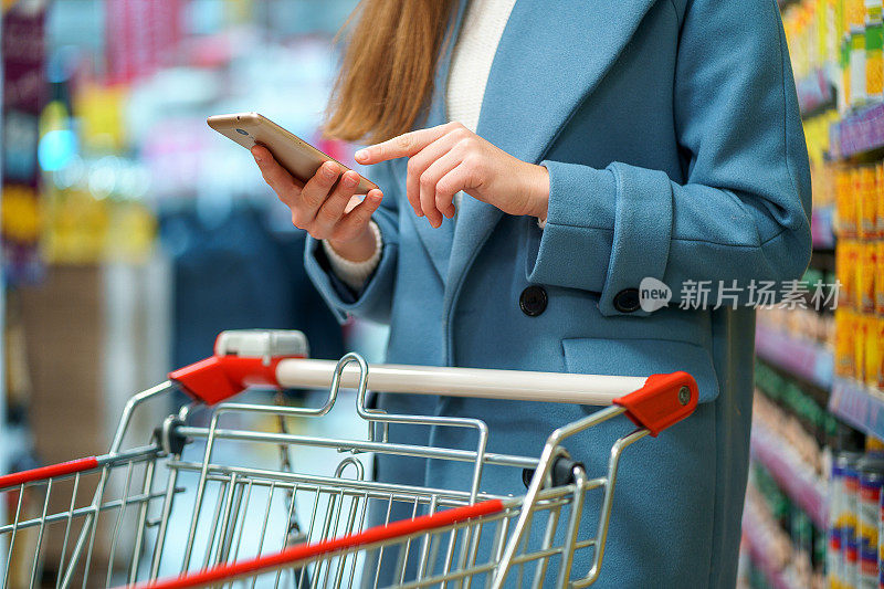 女买家推着购物车在商店过道上购物，智能手机上有购物清单