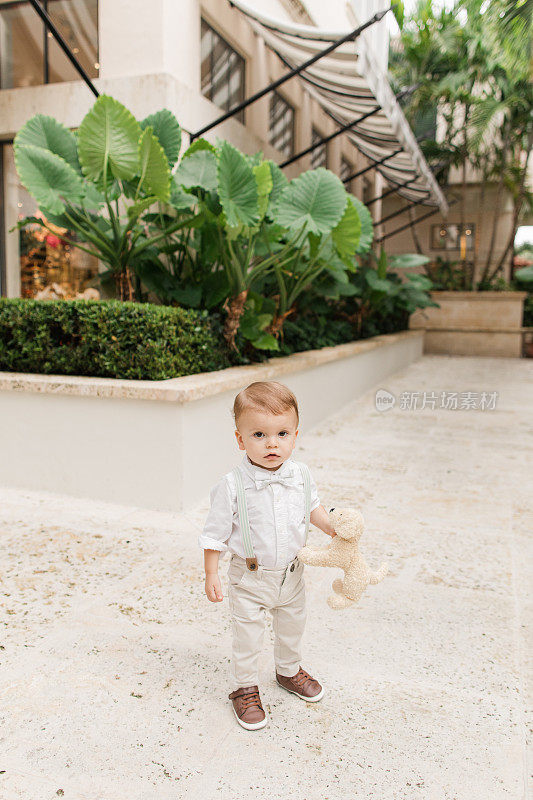 2023年5月，佛罗里达州热带棕榈滩，一个1岁的古巴裔美国小男孩，一头浅棕色短发，戴着鼠尾草绿色领结，穿着背带裤，白色纽扣衬衫，裤子和棕色鞋子，在花园里散步
