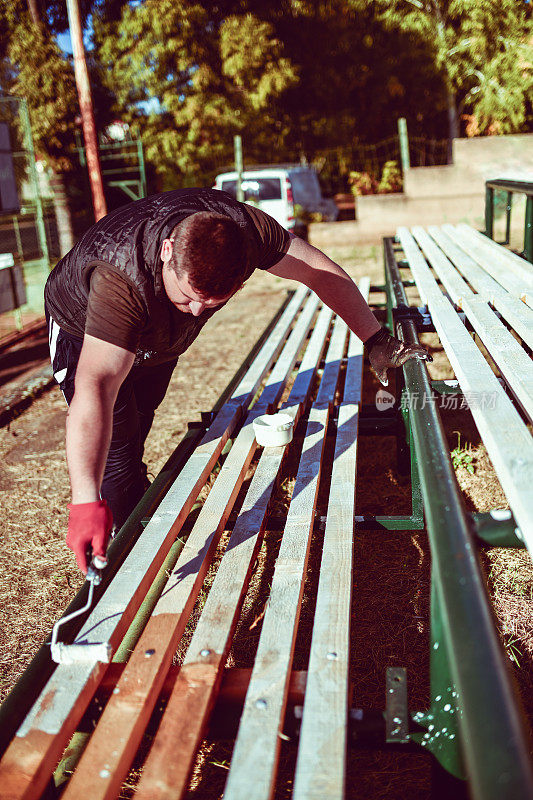 现代男性工人不知疲倦地在室外网球场的看台长凳上作画