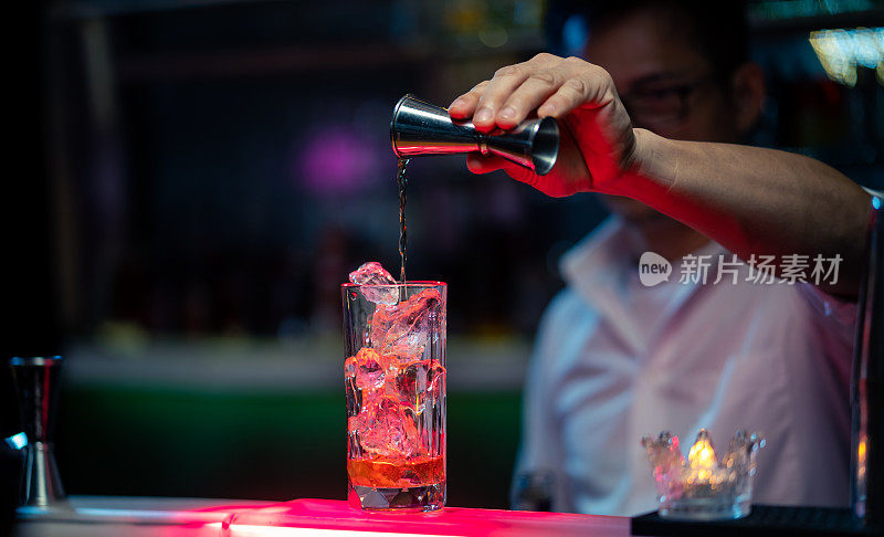 职业亚洲酒保，在吧台将酒倒入加冰块的长玻璃杯中，为夜总会服务。男调酒师调酒师准备酒精鸡尾酒饮料与烈酒架子的背景