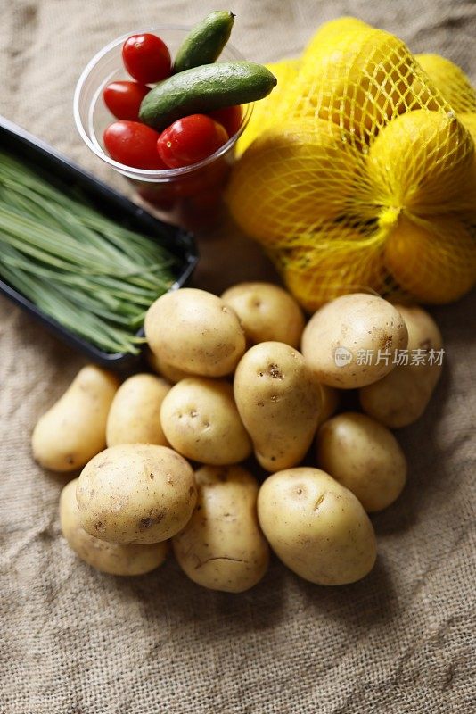 透明塑料杯里装着圣女果和小黄瓜，生土豆，黑色塑料盘子里放着细韭菜，粗麻布上的塑料网里放着柠檬