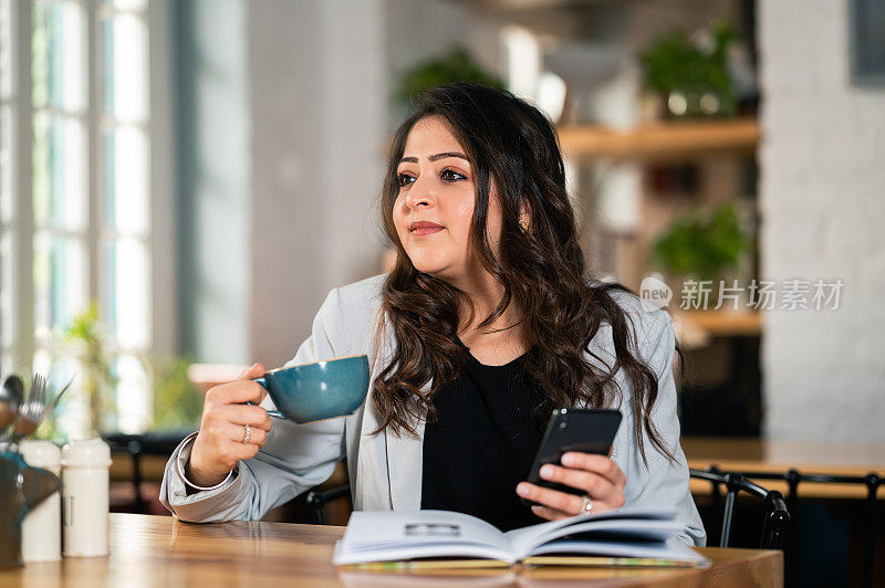 一名年轻女子一边使用智能手机，一边在咖啡馆看书、喝咖啡。