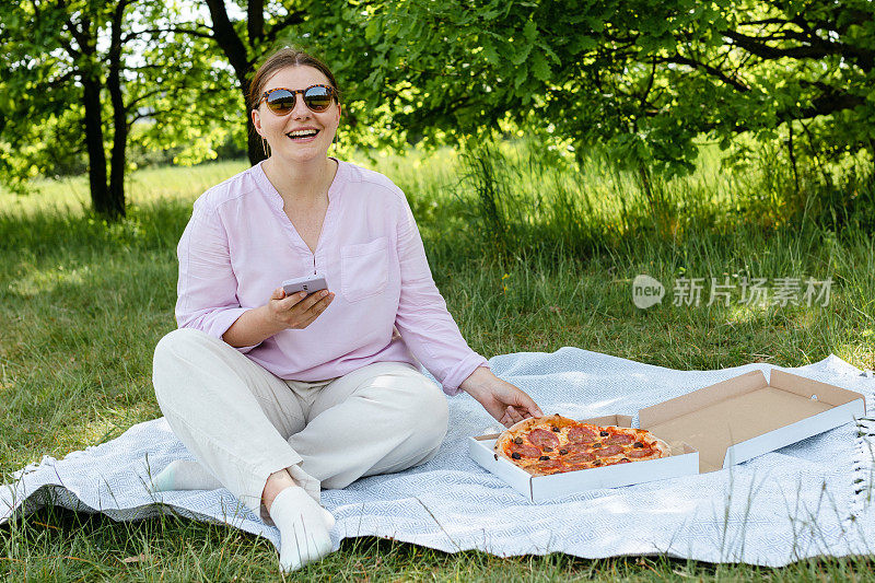 快乐的年轻女子吃披萨的自然背景和浏览互联网的电话。时尚美丽的女人肖像使用手机阅读短信户外。快餐或野餐横幅
