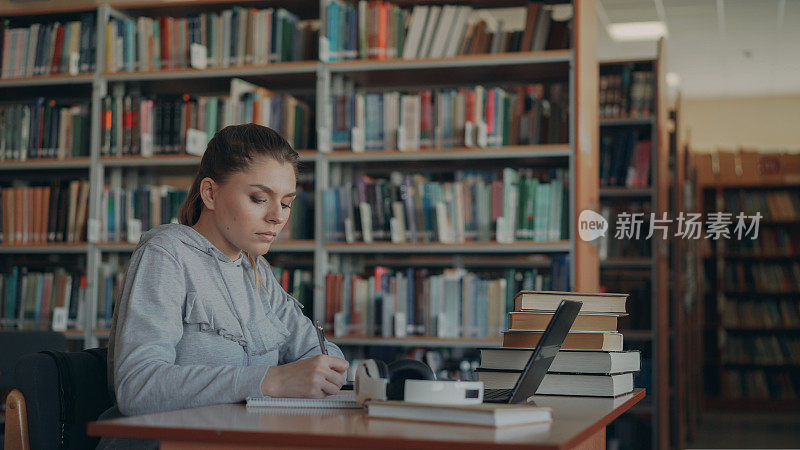 美丽严肃的白人女学生坐在图书馆的桌子上，用笔记本电脑在抄写本上写总结