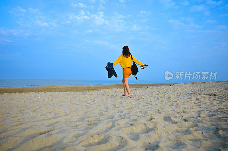 一个女人背着背包和鞋子走在秋天的海滩上的背影。