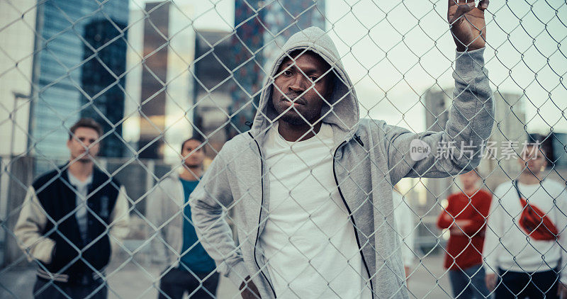 一个英俊的非洲裔美国人的肖像拿着栅栏，摆姿势，看着相机，而站在户外的城市位置。时尚多样的朋友站在他身后