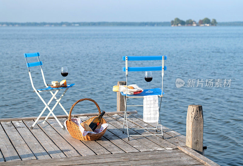 两张折叠椅，野餐篮，红酒，法棍面包和奶酪放在湖边的码头上