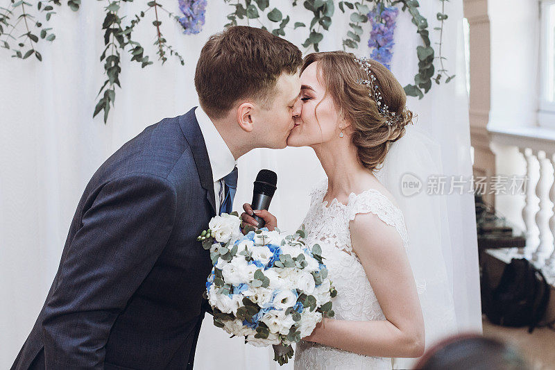 在婚宴上，新娘在祝酒后用麦克风与新郎接吻。奢华的紫色花朵装饰婚礼中心
