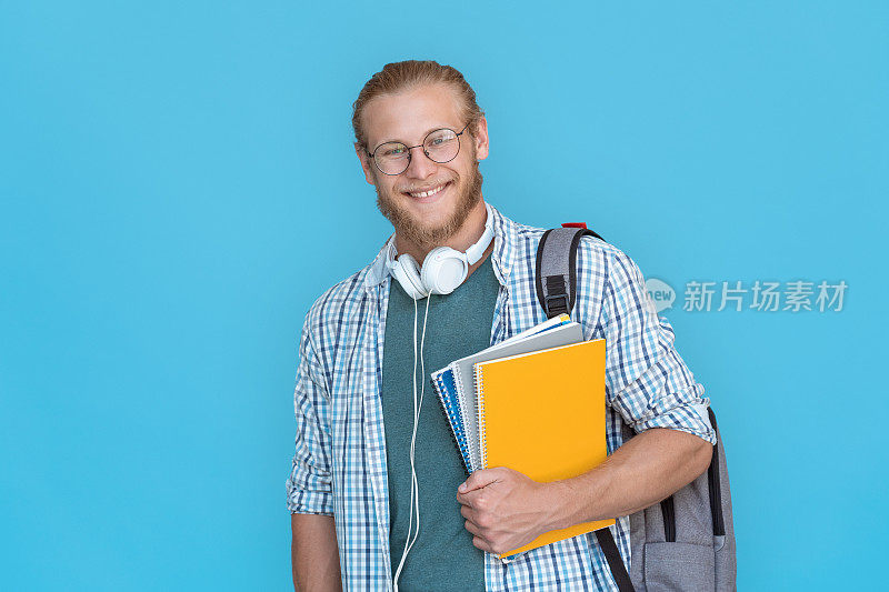 快乐自信的年轻大胡子帅哥学生拿着背包看练习册看着相机，随意的家伙大学老师站着用背包耳机隔离在蓝色工作室背景，肖像。