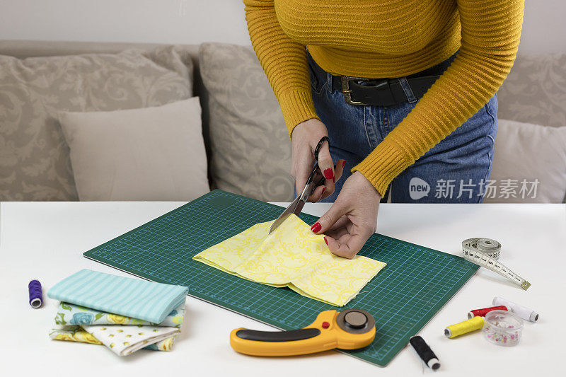 穿黄色毛衣的女人剪布料。桌子上有缝被子的工具。拼布刀、剪刀、衬里剪垫、自锁、线、卷尺。缝纫用品