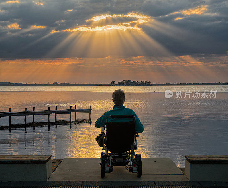 一位老人坐在轮椅上，在黄昏时分的湖面上沐浴着雄伟的阳光
