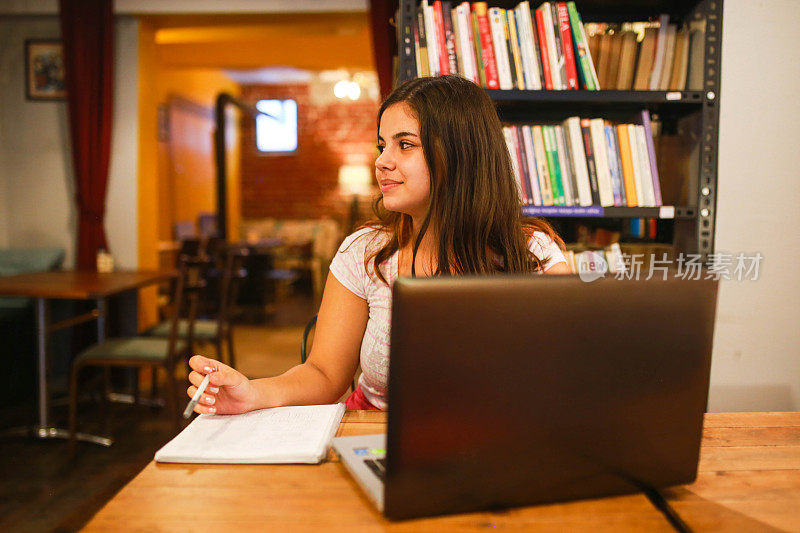 年轻女子拿着书在咖啡馆里用笔记本电脑学习。研究概念