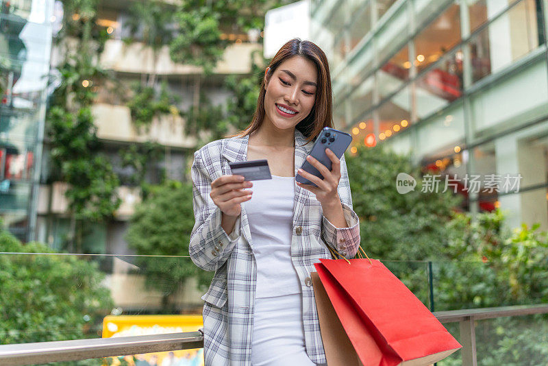 年轻的亚洲女商人管理信用卡在线交易，购物袋在手，无缝数字银行与智能手机随时随地