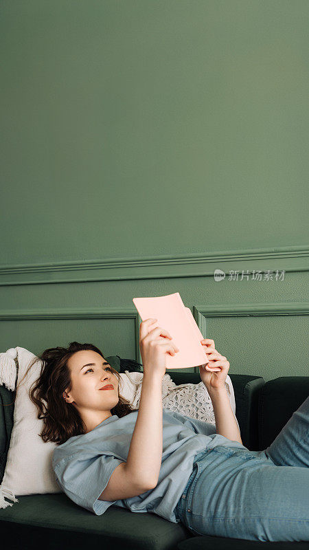 舒舒服服地躺在沙发上看书，享受休闲时光。在舒适的客厅里，微笑的年轻女士悠闲地阅读。家庭舒适中的放松与享受。