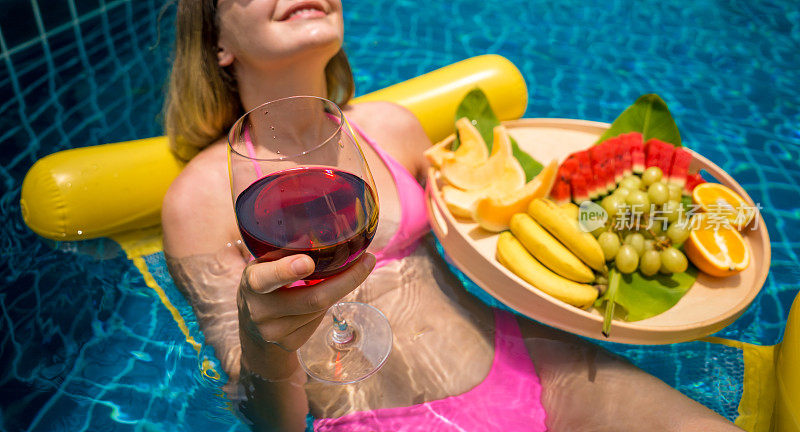 美丽的女人躺在漂浮的吊床上，在游泳池和酒和水果托盘