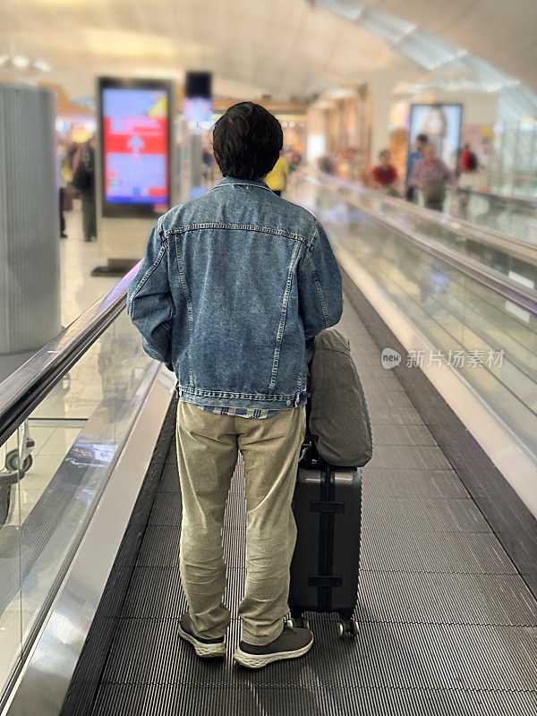 机场候机楼，印度旅客拿着飞机行李站在自动扶梯上旅行，行李堆在轮式行李箱上，焦点在前景，后视图