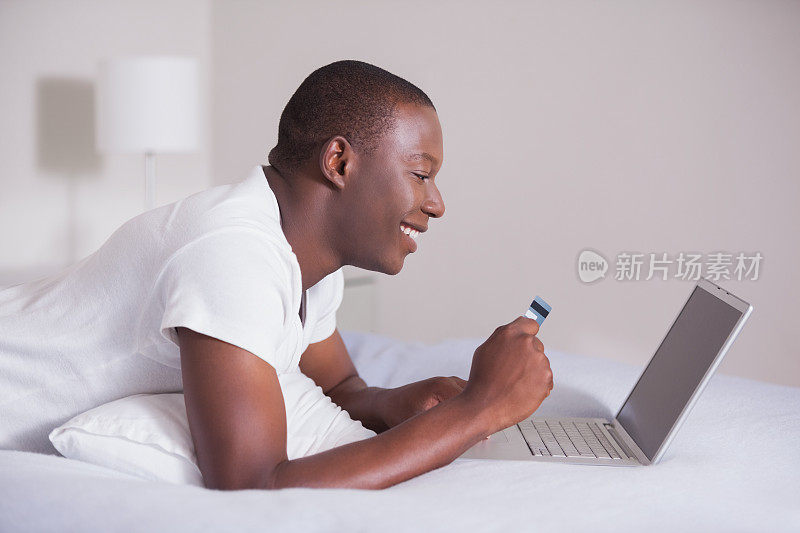 微笑的男人躺在床上用笔记本电脑在网上购物