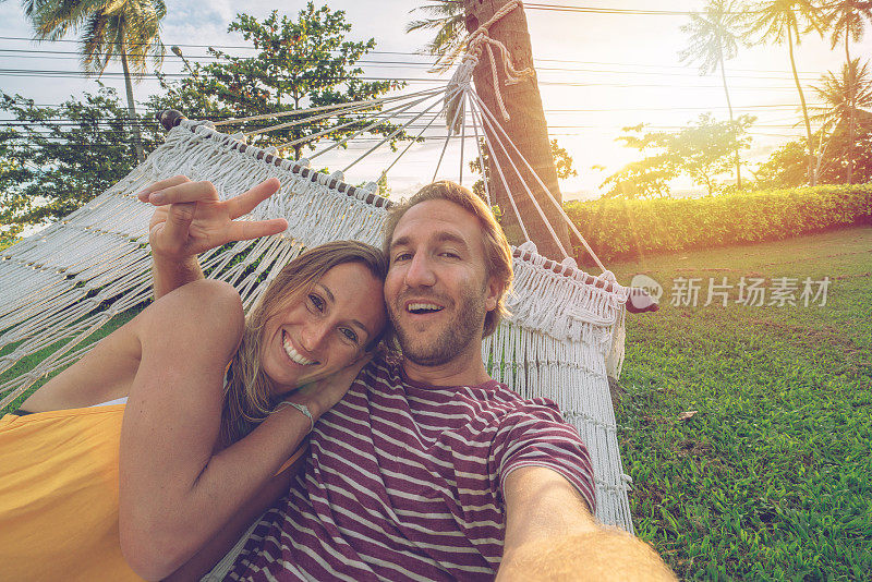 年轻夫妇在白色吊床上自拍，在热带气候下放松度假——人们在旅行中享受生活和自然，用移动设备技术捕捉凉爽的瞬间