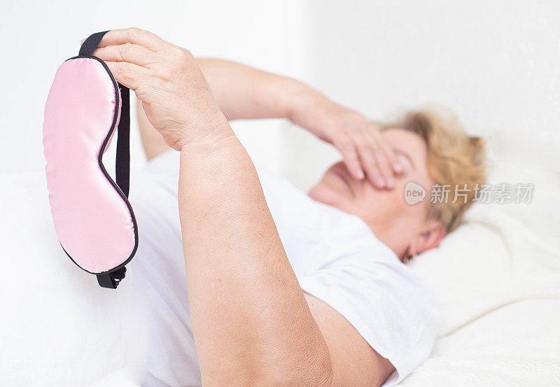 一位老年妇女在床上拿着一个睡眠面罩。失眠和镇静安眠药的概念