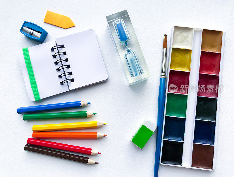 平铺文具学校和创意，绘画和工艺品(水彩画颜料，彩色铅笔，笔记本，沙漏)是在右边的白色纹理粗糙的水彩纸