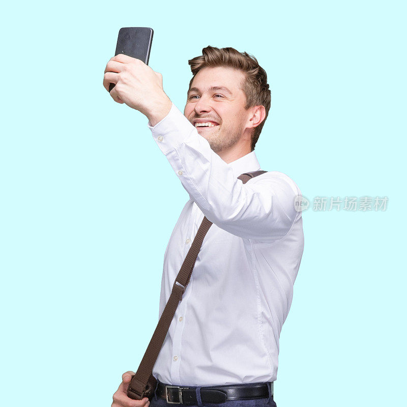 一名白种人青年男子站在蓝色背景前，戴着邮差包，拿着包，使用手机