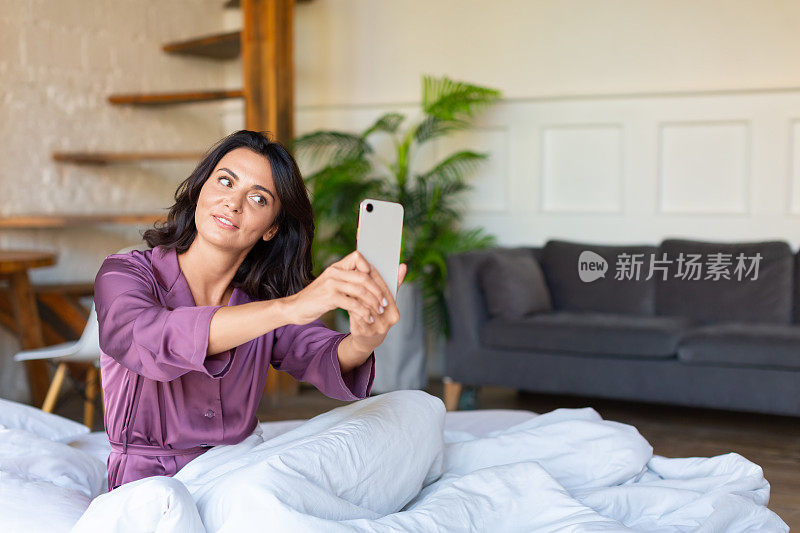 一位中年妇女躺在床上，手里拿着手机。她一觉醒来就在社交媒体上自拍。现代科技和数码成瘾的概念