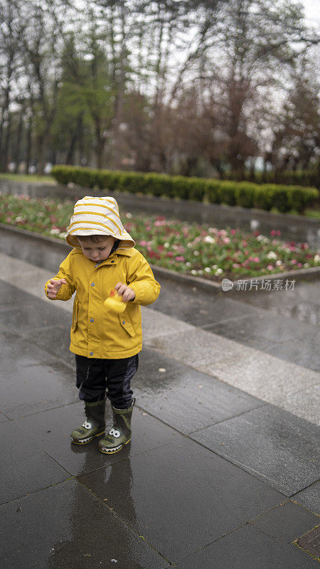 可爱调皮的小男孩，穿着亮黄色雨衣和胶靴，在春雨天潮湿的街道上和小水坑里的橡皮鸭玩耍。季节性天气步行概念