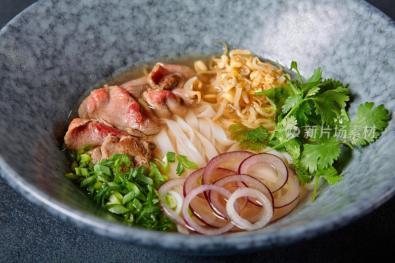 越南菜-牛肉米粉面汤或米粉汤。与新鲜的绿色蔬菜在蓝色碗上纹理深灰色的桌子。麻辣肉汤。