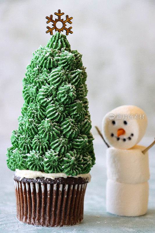 特写图像的个人，自制，圣诞树设计的巧克力纸杯蛋糕在纸蛋糕盒顶部的冰淇淋甜筒覆盖绿色黄油糖霜的管道，撒上糖霜，星星在上面，棉花糖雪人，大理石效果的背景