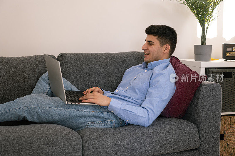 年轻英俊的男人坐在沙发上用笔记本电脑发短信