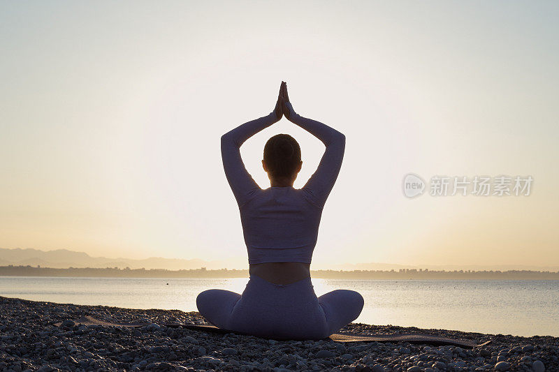 健康的年轻女子坐在瑜伽莲花姿势与闭上眼睛在大海前，在海滩上的日出冥想。