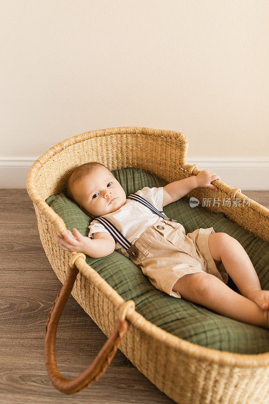 一个25周大的男婴穿着一件白色衬衫与海军条纹背带和卡其短裤，躺在一个舒适的绿色棉毯在海草摩西篮子周围的热带室内植物波西米亚热带氛围