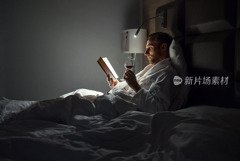 中年男子放松地躺在床上看书，拿着一杯红酒，床头灯开着。晚上放松，爱好，自由时间的概念。成年的概念。