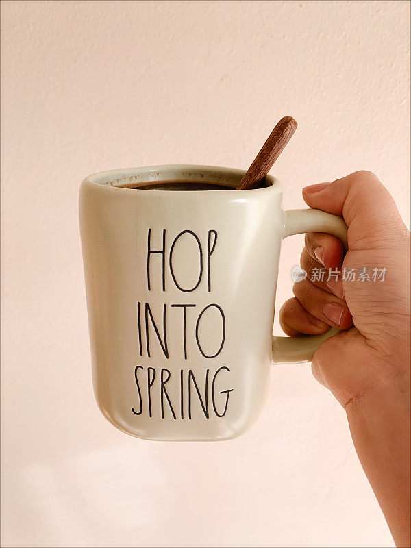 一个女人拿着一个浅绿色的杯子，上面写着“跳进春天”，里面装满了热黑咖啡和一个木制搅拌勺，简单的春天的乐趣，中性奶油色的背景与复制空间