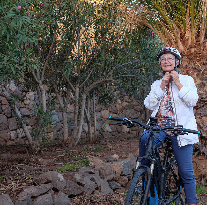 在户外活动中戴头盔骑电动自行车的运动老年妇女。老妇人在公园里骑自行车进行健康活动