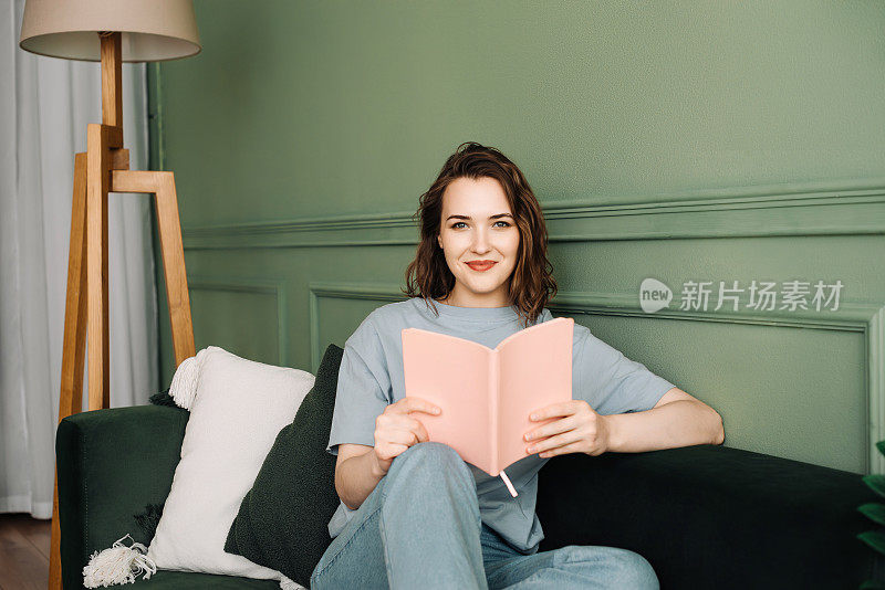 一个快乐的年轻女子舒舒服服地坐在沙发上看书，享受着一个放松的夜晚。放松的年轻女子坐在沙发上享受悠闲的晚间阅读。休息和放松
