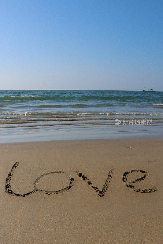 在阳光明媚的沙滩上用棍子写“爱”字的文字形象，水的边缘打破了海浪，在沙滩上柔软的金色沙滩上手写的字母，海边的海岸线，阳光明媚的蓝天，重点在前景，复制空间