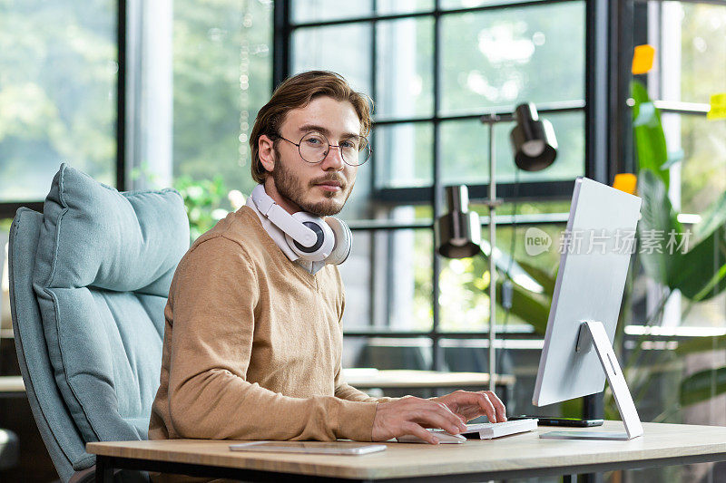 一个脖子上戴着耳机的严肃的办公室职员的肖像，坐在办公桌前，在电脑前工作，敲击键盘，严肃地看着相机