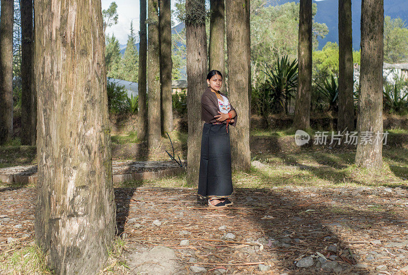 一位来自奥塔瓦洛的土著妇女走在她房子旁边的乡村小路上，她穿着传统的服装，看着镜头