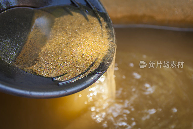 特写的黄金淘金锅与筛砂。景深较浅，聚焦于沙子从锅边流入水中。