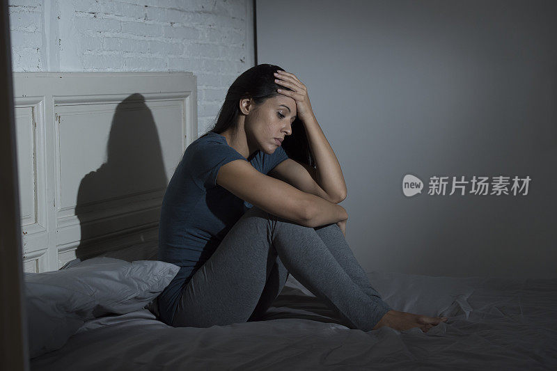 年轻美丽的悲伤和沮丧的拉丁女人坐在家里卧室的床上，看起来沮丧和担心遭受抑郁和头痛