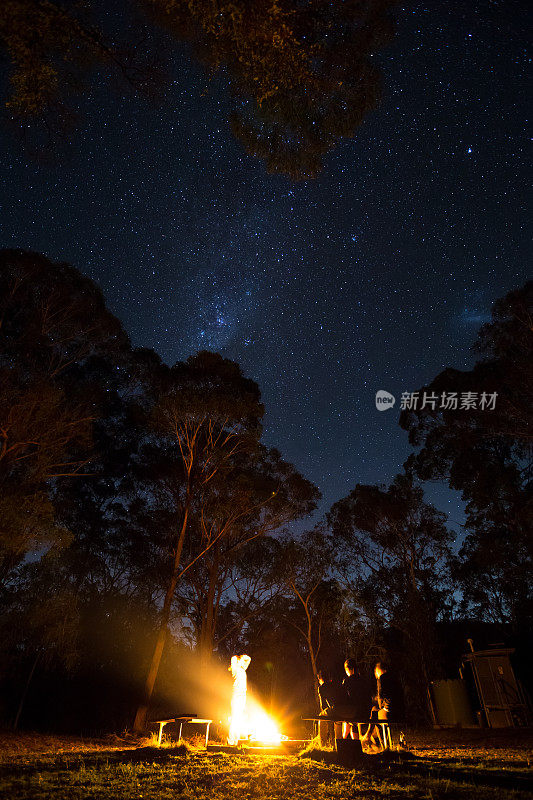 一群露营者围着篝火站着，上面闪烁着明亮的星星