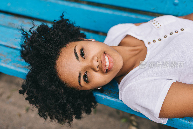 肖像快乐的非洲裔美国妇女休息在户外长椅上的公园，微笑与牙齿的微笑，并看着镜头。美丽年轻的黑皮肤女性享受健康的生活。人
