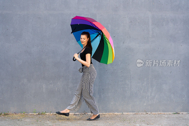 一名年轻女子撑着彩色雨伞走在一堵混凝土墙前