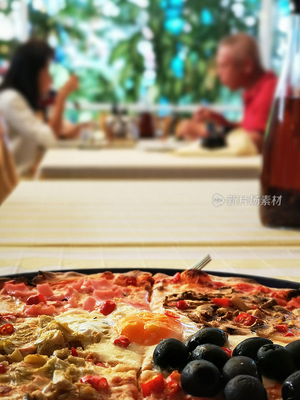 四个阶段的美味正宗意大利披萨中间放一个鸡蛋和新鲜的辣辣椒，在传统的披萨烤箱里烤好，放在餐桌上，做成一道菜。背景中可以看到两个人和一个人