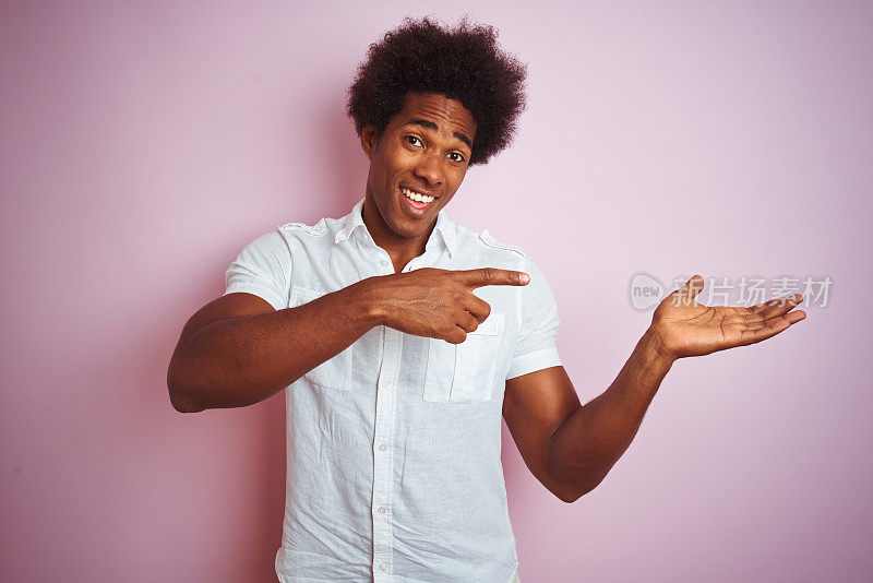一个留着非洲式发型、身穿白衬衫的年轻美国男子站在孤立的粉红色背景下，对着镜头微笑着，用手指着镜头。