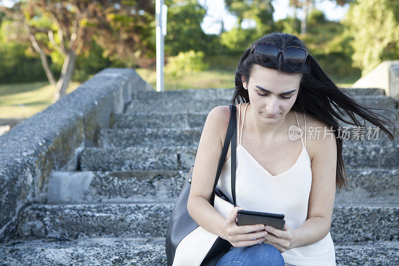 一个十几岁的女孩坐在水泥台阶上，手里拿着电子阅读器