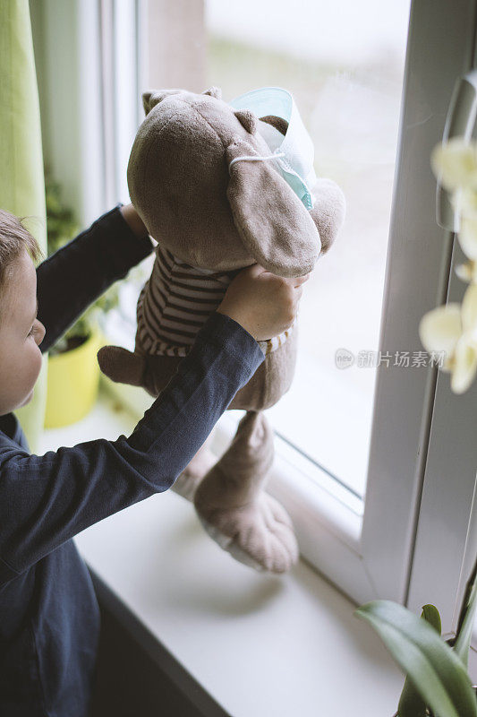小男孩戴着防护口罩，带着闷热的玩具，从窗户往外看。预防传染病