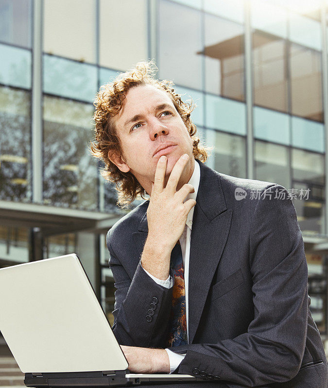 一位西装革履的商人坐在现代化的玻璃办公大楼外，带着他的笔记本电脑，若有所思