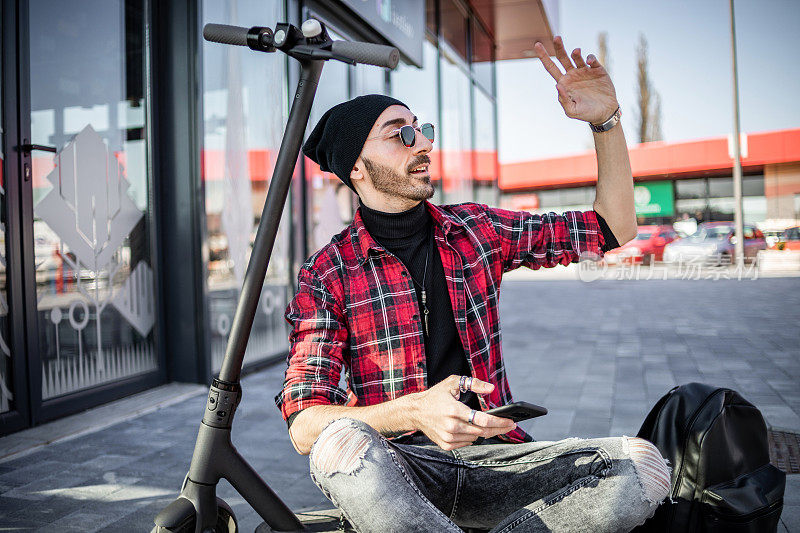 一个骑着电动滑板车的潮人，他坐在滑板车旁边，用着智能手机。技术、生态的移动性概念。